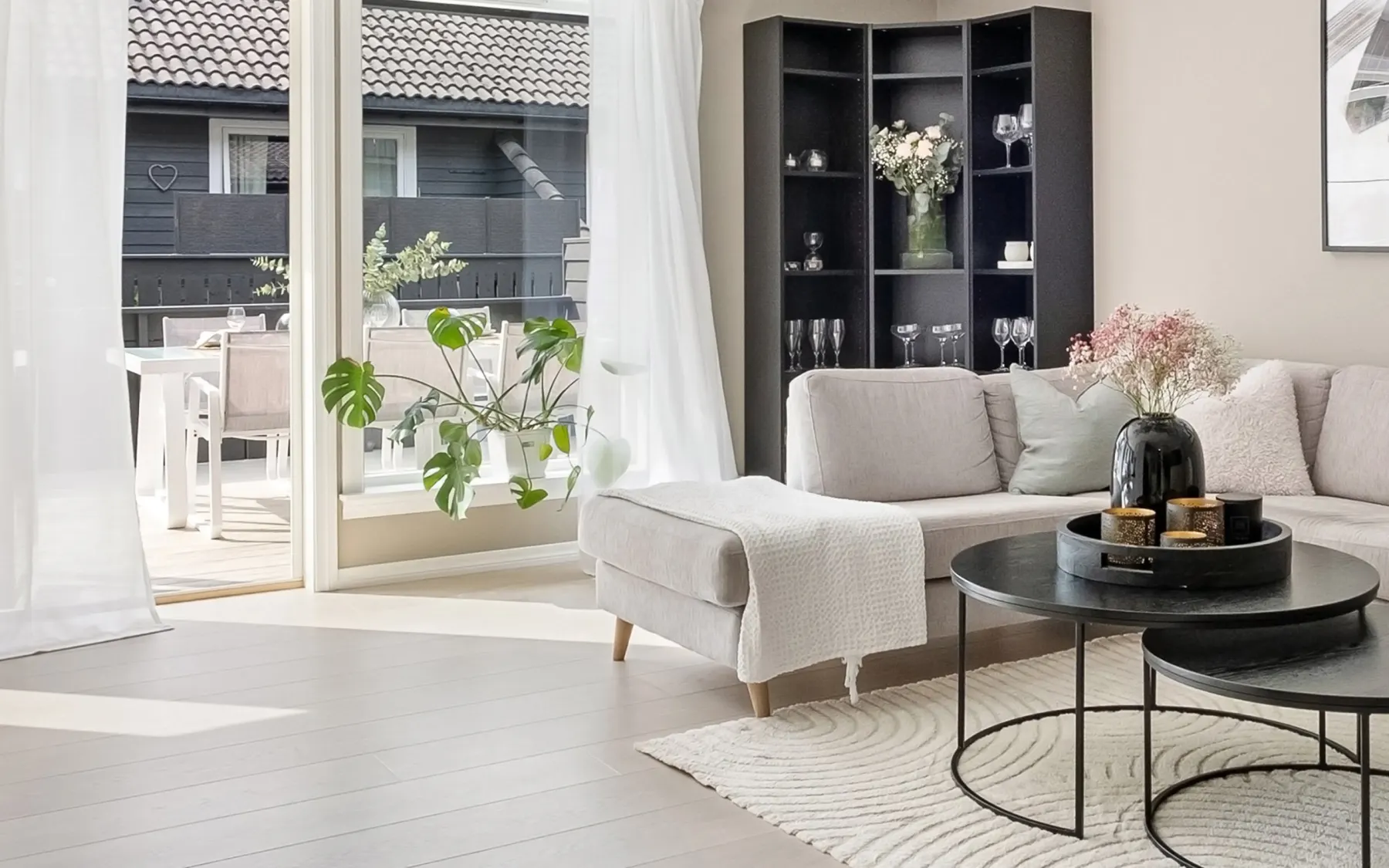 Modernt vardagsrum med grå soffa, vita kuddar, svarta soffbord, växter och glasdörrar mot uteplats.