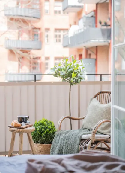 Liten balkong med en rottingstol, gröna kuddar och en filt. Bredvid finns ett litet bord med en kopp kaffe och croissant. Växter och en utsikt över flerbostadshus i bakgrunden.